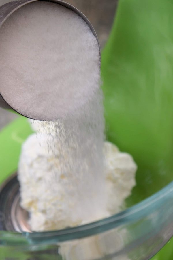 Picture of sugar being poured over cream cheese in a large glass bowl.