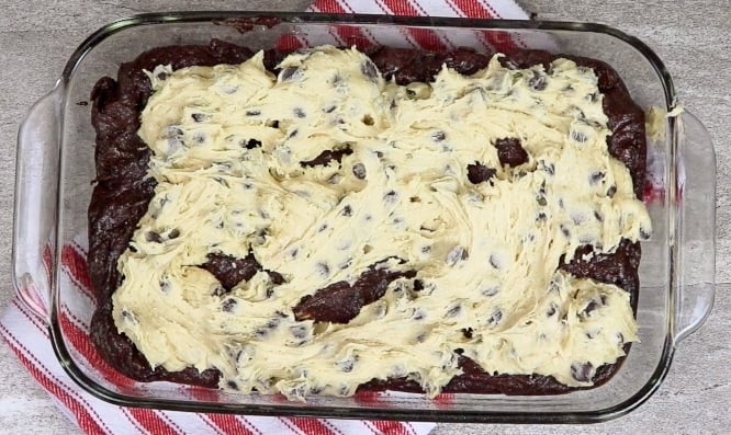 Cookie dough spread out on top of brownie batter in a glass baking dish. 