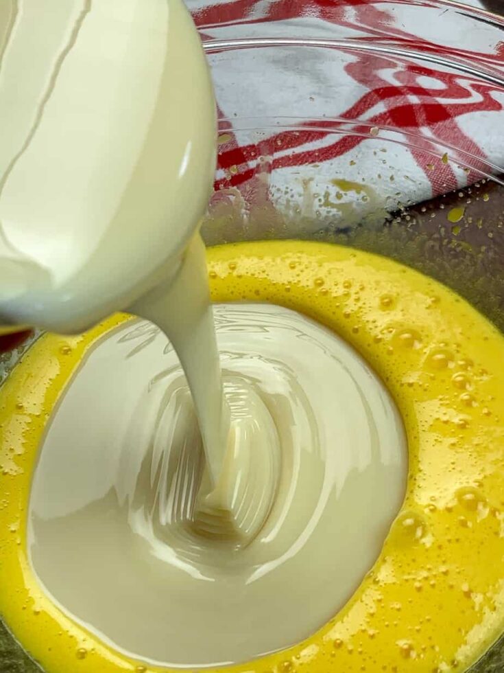 Pouring sweetened condensed milk into a glass bowl with egg yolks