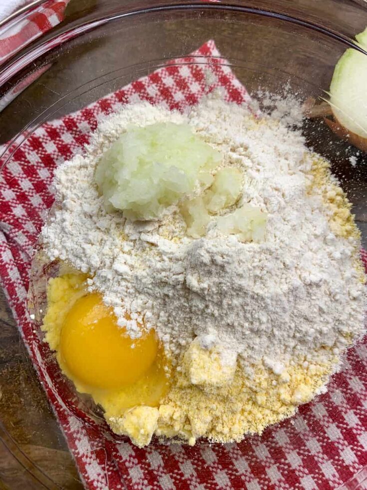 Diced onions, cornbread mix, flour, and an egg in a glass bowl.