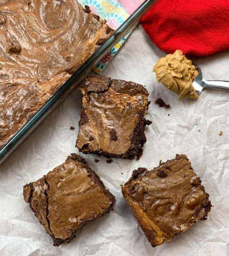 Three brownies sitting next to a pan of brownies and a spoonful of peanut butter